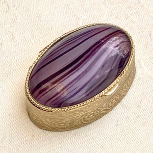 Può includere: Scatola portagioie ovale con base in metallo color oro e top in pietra a strisce viola e nere. La scatola ha un motivo floreale decorativo sui lati e un bordo a forma di corda attorno alla pietra.