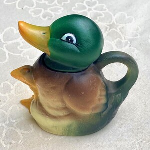 Pode incluir: Bule de cerâmica em forma de pato. O pato tem cabeça verde e bico amarelo. O corpo é castanho e bege, com uma pega verde. O olho do pato é branco com uma pupila preta. Artigo decorativo.