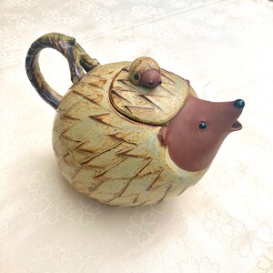 Op de afbeelding: Een keramische theepot in de vorm van een egel, met een bruin gezicht en een kleine kever op het deksel. De theepot heeft een getextureerd, lichtgroen en bruin lichaam met een takvormige handgreep.