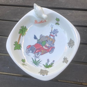 Pode incluir: Prato de cerâmica branca com uma pega em forma de pássaro. O prato apresenta uma ilustração de Babar com um carro vermelho e palmeiras. A palavra "Babar" está escrita em vermelho. O prato tem uma forma quadrada com bordas arredondadas.