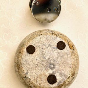 May include: A silver clock face sits above a rounded, stone-like object. The stone has a mottled beige and grey appearance with three dark brown, circular pads on its underside. The clock face has a shiny, reflective surface.