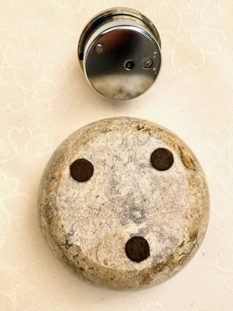 May include: A silver clock face sits above a rounded, stone-like object. The stone has a mottled beige and grey appearance with three dark brown, circular pads on its underside. The clock face has a shiny, reflective surface.
