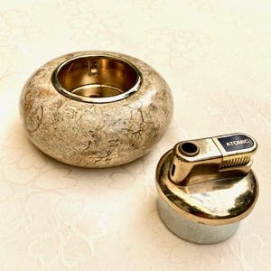 May include: A beige, round candle holder with a gold interior and a gold lighter with the word "Atomic". The candle holder has a speckled pattern, and the lighter has a silver base.