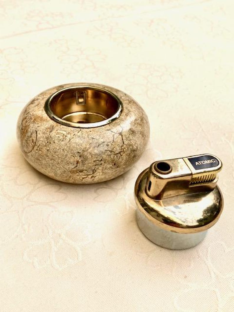 May include: A beige, round candle holder with a gold interior and a gold lighter with the word "Atomic". The candle holder has a speckled pattern, and the lighter has a silver base.