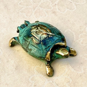 Op de afbeelding: Kleine bronzen schildpadfiguur. De schild heeft een groen patina, met goudkleurige accenten op de vinnen en kop. Een decoratief item, waarschijnlijk voor decoratie.