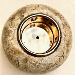 May include: A circular candle holder made of stone-like material, featuring a gold-coloured metal insert. The exterior has a mottled beige and grey appearance with a rough texture. The metal insert is circular.