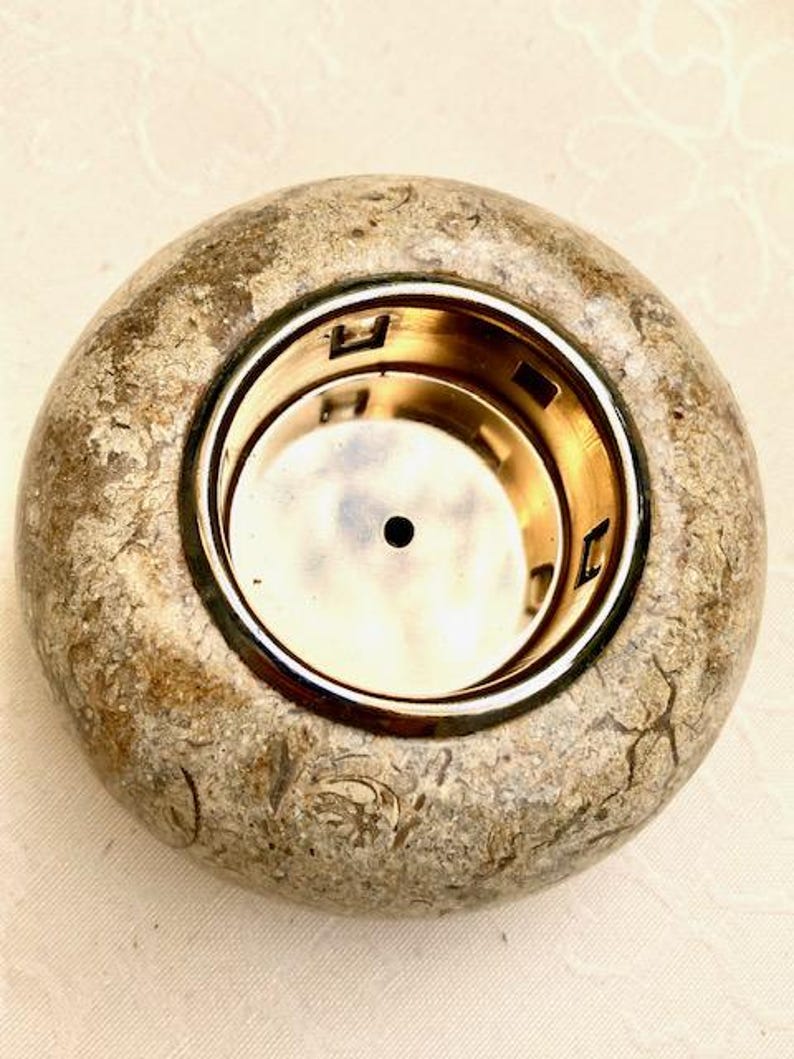 May include: A circular candle holder made of stone-like material, featuring a gold-coloured metal insert. The exterior has a mottled beige and grey appearance with a rough texture. The metal insert is circular.