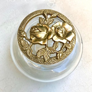 May include: A clear glass jar with a decorative brass lid featuring two roses and leaves. The lid has a textured border and a circular design. The jar is round and has a pearlescent sheen.