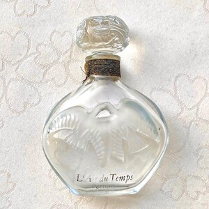 Può includere: Un flacone di profumo in vetro trasparente con un tappo decorativo. La bottiglia ha un design smerigliato di colombe e le parole "L'Air du Temps" in scrittura nera. La bottiglia è alta circa 10 cm.