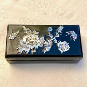 Peut inclure: Une boîte noire rectangulaire avec une finition brillante. Le couvercle est orné d'un motif floral incrusté de nacre irisée. Les fleurs et les feuilles sont détaillées, créant un objet de rangement décoratif.