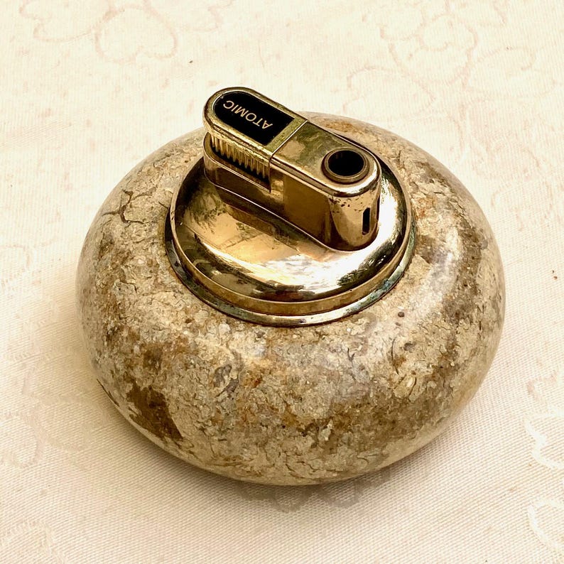 May include: A vintage lighter with a gold-coloured metal top and the word "ATOMIC" printed on it. The lighter is set on a round, stone-like base with a mottled, beige and brown pattern. The lighter's design suggests a retro aesthetic.