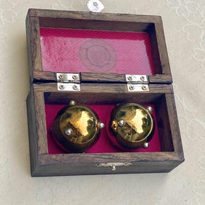 Op de afbeelding: Een houten kist met een scharnierend deksel, gevoerd met rode stof, herbergt twee goudkleurige, bolvormige objecten. De kist is gemaakt van donker hout en heeft zilverkleurige scharnieren en een sluiting. De bolvormige objecten hebben kleine zilveren accenten.