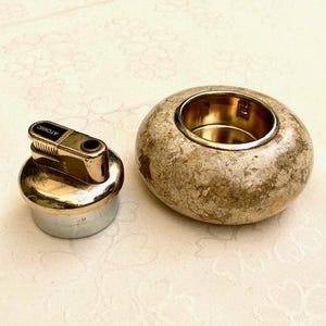 May include: A gold-toned lighter with the word "ATOMIC" printed on it, next to a round, speckled stone ashtray with a gold-coloured interior. The lighter has a silver base. The ashtray is a light brown colour.