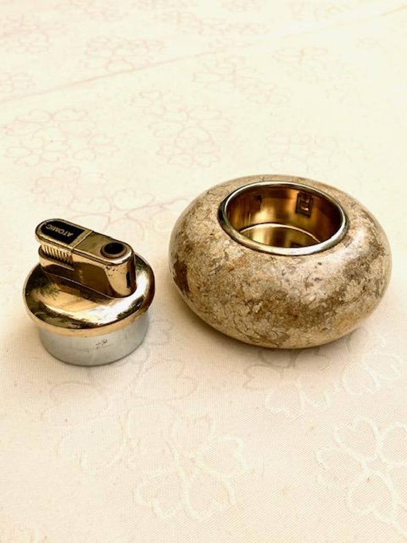 May include: A gold-toned lighter with the word "ATOMIC" printed on it, next to a round, speckled stone ashtray with a gold-coloured interior. The lighter has a silver base. The ashtray is a light brown colour.