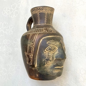 Può includere: Un vaso in ceramica marrone scuro a forma di testa umana con un solo manico. La superficie presenta incisioni a linee chiare che raffigurano tratti del volto stilizzati, motivi geometrici sul collo e disegni di animali sul lato.