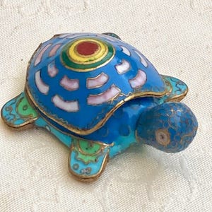 Pode incluir: Estatueta decorativa de tartaruga com carapaça azul, apresentando um design central de círculos vermelhos, amarelos e verdes. A carapaça da tartaruga tem detalhes brancos e rosa, com acabamentos dourados. Um item decorativo único.