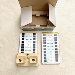 Puede incluir: Un visor estereoscópico de plástico beige con una palanca de enfoque negra, acompañado de dos tarjetas rectangulares con múltiples diapositivas y una caja de cartón etiquetada STEREOSCOPE VIEWER THREE-D, con imágenes del Mont Saint-Michel.