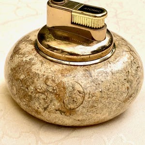 May include: A vintage lighter with a gold-toned metal top and the word "ATOMIC" printed on it. The base is a rounded, speckled stone in shades of beige and brown. A collectible lighter.