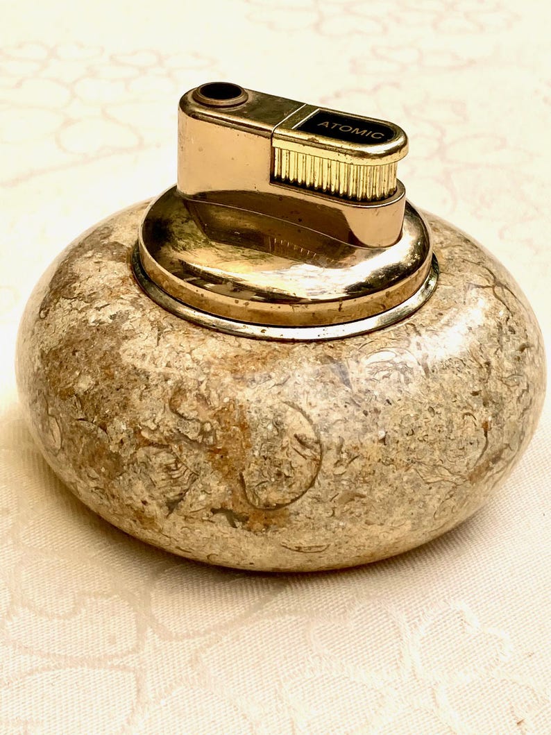 May include: A vintage lighter with a gold-toned metal top and the word "ATOMIC" printed on it. The base is a rounded, speckled stone in shades of beige and brown. A collectible lighter.