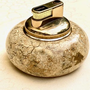 May include: A vintage lighter with a gold-toned metal top and a textured, stone-like base. The top is marked "ATOMIC". The lighter has a round, pebble-like shape, and the base has a mottled, earthy colour scheme.