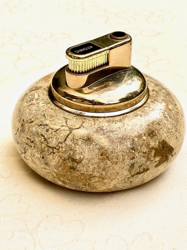 May include: A vintage lighter with a gold-toned metal top and a textured, stone-like base. The top is marked "ATOMIC". The lighter has a round, pebble-like shape, and the base has a mottled, earthy colour scheme.