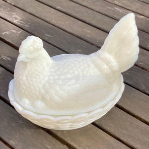 Puede incluir: Un plato cubierto en forma de gallina, blanco y opalino. La gallina se asienta sobre una base de tejido de cesta, con cabeza y plumas de cola detalladas. El plato es de vidrio y probablemente vintage.