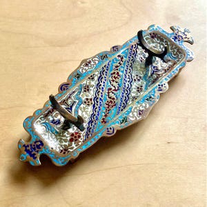 Op de afbeelding: Een sierlijk, rechthoekig sieradenbakje met een gedetailleerd emaille ontwerp. Het bakje heeft een goudkleurig metalen frame met een turquoise, blauw, rood en wit bloemmotief. Het heeft twee metalen handvatten en decoratieve uiteinden.