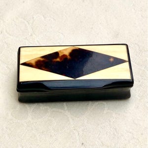Puede incluir: Caja rectangular negra con tapa color crema y diseño central de rombo en carey y azul oscuro. La caja tiene un acabado brillante y un diseño sencillo y elegante, adecuado para guardar objetos pequeños.