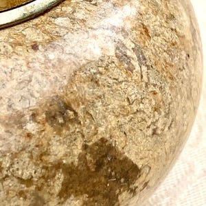 May include: Close-up of a decorative, round object with a textured, mottled surface in shades of beige, brown, and tan. A silver-coloured metal band encircles the top. The object has a rustic, aged appearance.