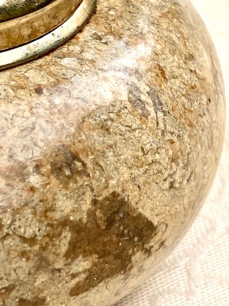 May include: Close-up of a decorative, round object with a textured, mottled surface in shades of beige, brown, and tan. A silver-coloured metal band encircles the top. The object has a rustic, aged appearance.
