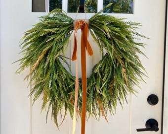 May include: A circular wreath constructed from dense, feathery green foliage with subtle hints of yellow and brown. The top of the wreath is bound with twine and features two long, hanging ribbons in a solid burnt orange velvet fabric.