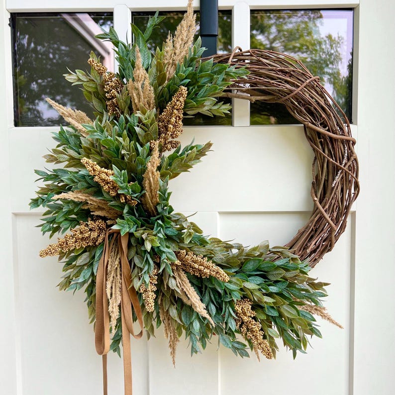 May include: A brown grapevine wreath with a brown velvet ribbon and green and gold foliage. The wreath is decorated with dried grasses and small gold berries.