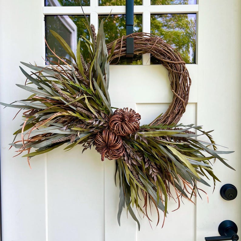 May include: A grapevine wreath with a copper colored floral accent and green and brown foliage. The wreath is hanging on a white door.