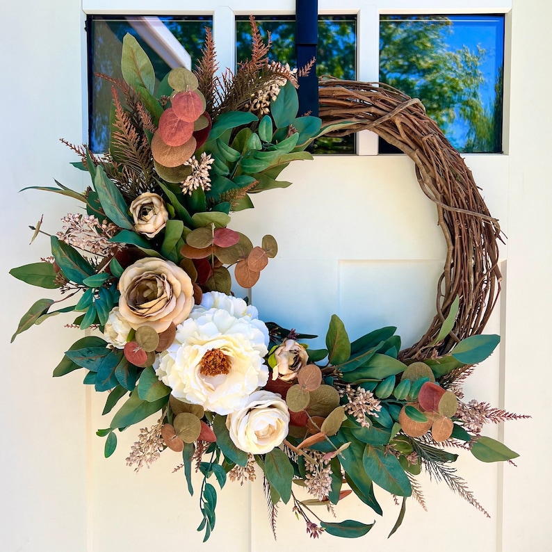 May include: A brown grapevine wreath decorated with artificial flowers and greenery. The wreath features white, cream, and brown flowers, along with green leaves and sprigs of brown foliage.