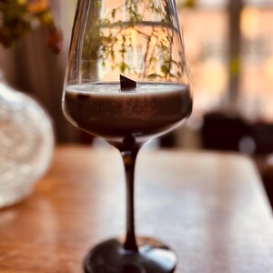 Puede incluir: Una copa de vino transparente con tallo y base oscuros, llena de un líquido cremoso marrón. Un pequeño objeto oscuro flota en el líquido. La copa está sobre una superficie de madera, con un fondo borroso de ventana y follaje.