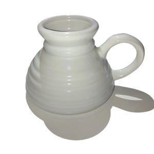 May include: A cream-colored ceramic mug with a handle. The mug has a rounded base and a flared top. The body of the mug features a textured, ridged design. The handle is a simple loop shape.