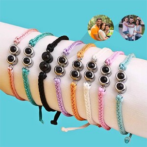 May include: A selection of colourful personalized bracelets, each with three black stones set in silver-tone metal. The bracelets are displayed on a wrist, showing the braided cord and adjustable design. Family photos are in the background.