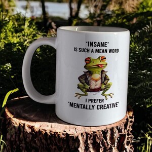 May include: White ceramic mug featuring a green frog in a leather jacket and sunglasses. The mug has the text "'INSANE' IS SUCH A MEAN WORD I PREFER 'MENTALLY CREATIVE'" printed on it.