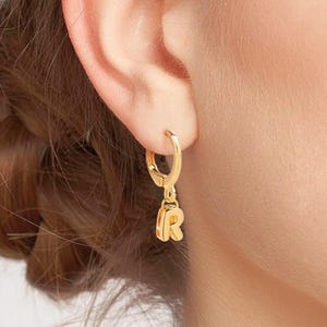 Może przedstawiać: Złoty kolczyk w kształcie koła z wiszącym wisiorkiem z literą "R". Kolczyk jest przymocowany do ucha. Biżuteria ma prosty, elegancki design. Kolczyk to mały, delikatny element biżuterii.