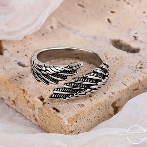 May include: A silver ring with a wing design. The ring has detailed feather patterns, with the wings wrapping around the band. It is displayed on a textured, light brown surface.
