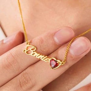 May include: A gold-colored necklace with the name "Bonnie" in cursive script and a heart-shaped red gemstone. The necklace is on a delicate chain, and the gemstone is set in a gold bezel.