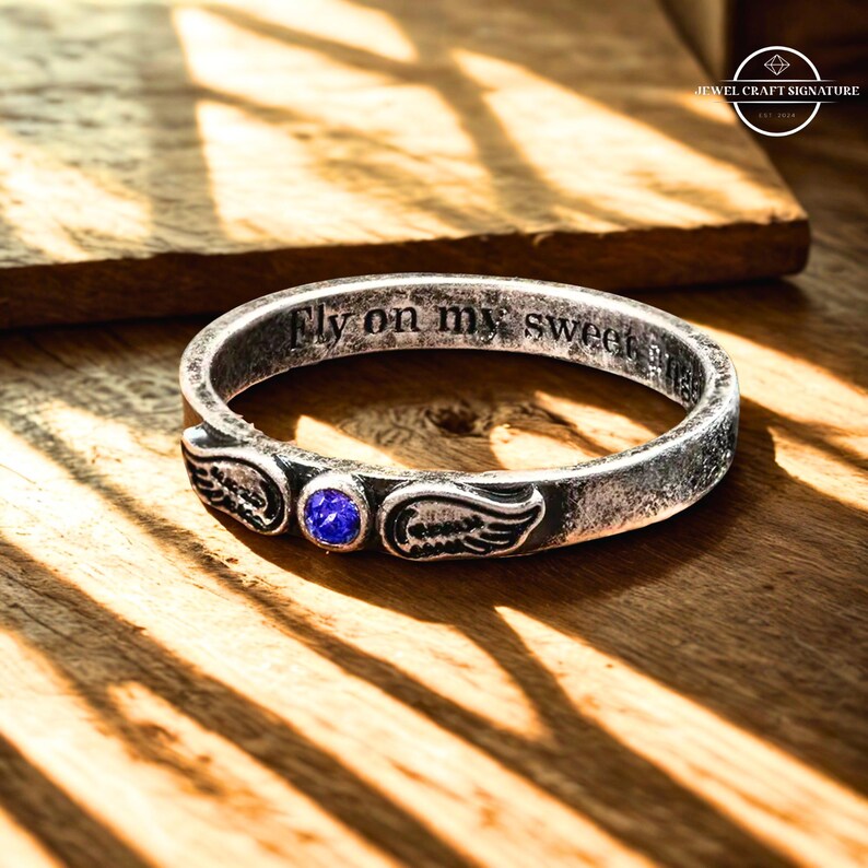 K&ouml;nnte beinhalten: Ein silberner Ring mit der Inschrift "Fly on my sweet" und Fl&uuml;gelmotiven. Der Ring hat einen blauen Edelstein und liegt auf einer Holzoberfl&auml;che. Der Ring ist ein Schmuckst&uuml;ck.