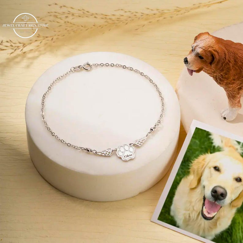 May include: A silver charm bracelet with a paw print charm with wings and the name "Olivia" engraved on it. The bracelet is on a white surface with a photo of a golden retriever in the background.