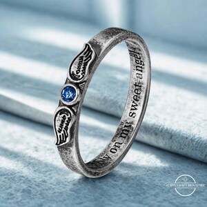 K&ouml;nnte beinhalten: Silberner Ring mit Engelsfl&uuml;gel-Design und blauem Edelstein. Der Ring tr&auml;gt die Inschrift "fly on my sweet angel". Der Ring liegt auf einer hellblauen Oberfl&auml;che.