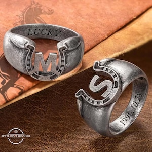 May include: Two silver-toned rings with horseshoe designs. One ring has the letter "M" in the center and the word "LUCKY" engraved on the band. The other ring has the letter "S" and the date "1990.10.28" engraved on the band. The rings are on a brown surface.