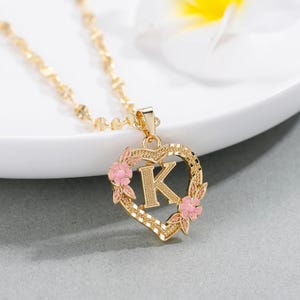 May include: Gold necklace with a heart-shaped pendant featuring the letter K surrounded by pink flowers.