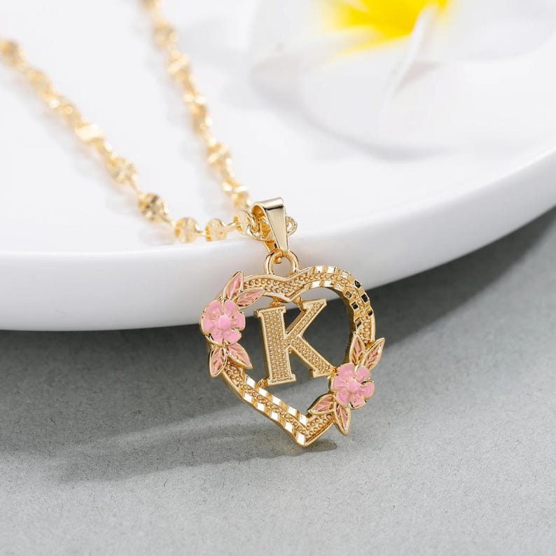 May include: Gold necklace with a heart-shaped pendant featuring the letter K surrounded by pink flowers.