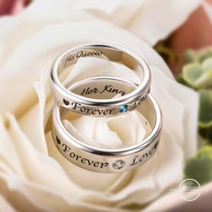 Könnte beinhalten: Zwei silberne Ringe mit den Inschriften "His Queen" und "Her King" liegen auf einer weißen Rose. Die Ringe tragen das Wort "Forever" und ein kleines Herz. Ein Ring hat einen blauen Edelstein, der andere einen klaren Edelstein.