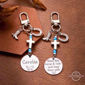 Puede incluir: Dos llaveros plateados con dijes de bota de vaquero, herradura y cruz. Uno tiene el nombre "Cerelia" y un gráfico de caballo. El otro dice "Bless this horse & rider and keep them safe."