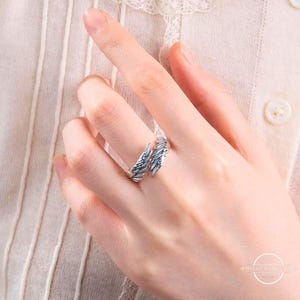 May include: A silver-colored ring with a feather design, worn on a finger. The ring wraps around the finger with two detailed feather elements. The ring is shown on a hand with a light-colored background.
